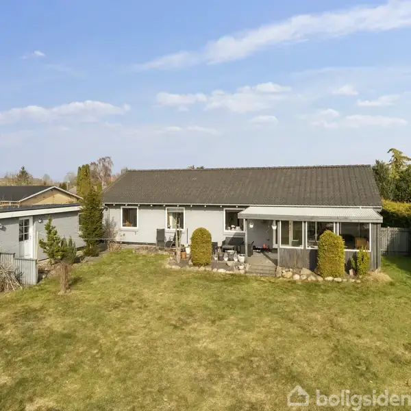 Et lille, hvidt hus med fladt tag står i en have med velplejet græs og buske. Ved huset er en overdækket terrasse med havemøbler. Skyfri blå himmel i baggrunden.