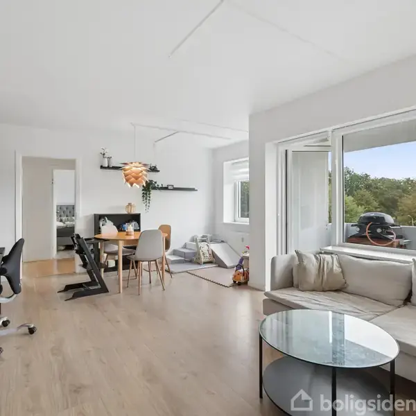Sofa står i en lys stue foran et stort vindue, der åbner ud til en altan. Ved siden af er der spisebord med stole. Møbleret med skab og hylder.