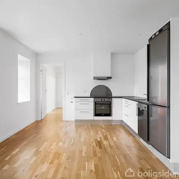 Køkken med hvide skabe og sorte apparater som køleskab og ovn, placeret på trægulv. Rummet er lyst med store vinduer, og en dør fører til et tilstødende værelse.