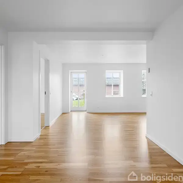 Et rum med trægulv uden møbler. Lyse vægge og to vinduer i baggrunden giver naturligt lys. Flere åbninger leder til tilstødende rum.