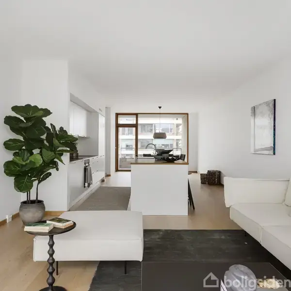 En moderne stue med en hvid sofa, sofabord og grøn plante. Ovenlys fra store vinduer bagest i rummet. Lyst trægulv og minimalistisk indretning skaber en åben og lys atmosfære.