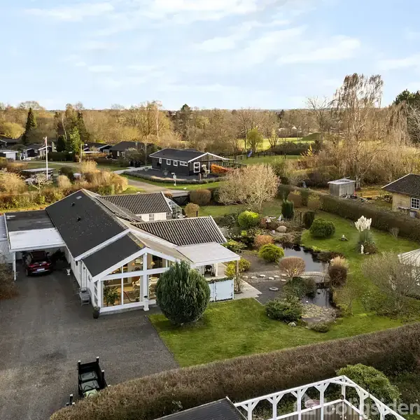 Et moderne hus med sort tag ligger centralt omgivet af en velplejet have med buske og træer. I baggrunden ses flere huse og en skovbevokset horisont.