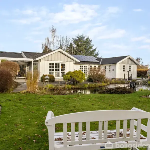 Hvid bænk står i en have foran et hvidt hus med solpaneler på taget. Haven indeholder en dam, buske og træer. Himlen er blå med skyer.
