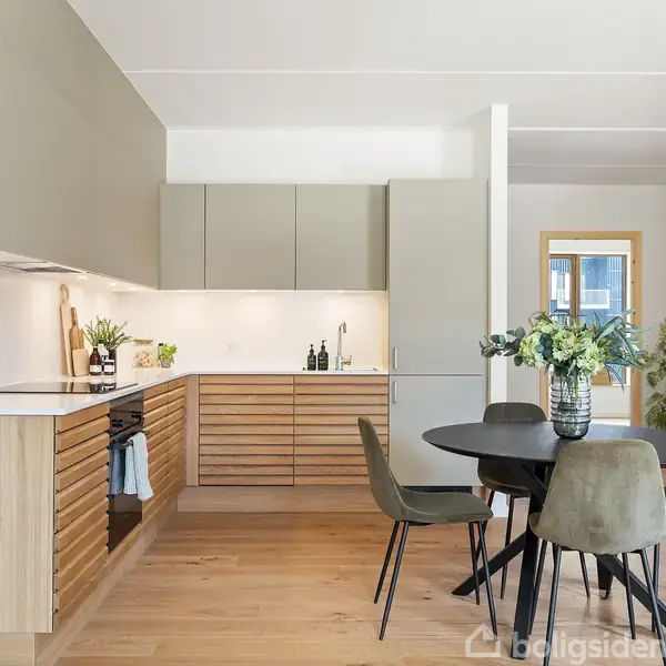 Moderne køkken i åben stil med træskabe og hvide flader. Rundt spisebord med fire stole stående i spisestue området, prydet med en plantevase. Lys træparketgulv rundt omkring.