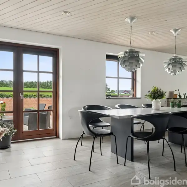 Et moderne køkken med stort spisebord og seks sorte stole står ved en glasdør, der fører ud til en veranda. Flere planter dekorerer rummet.