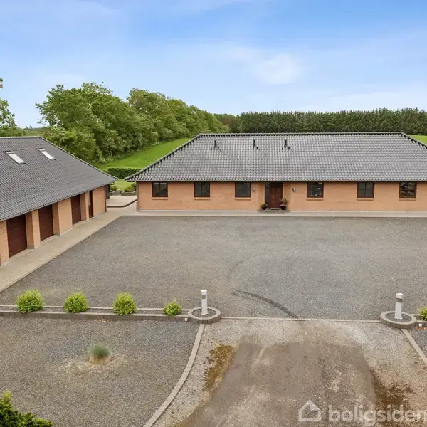 En stor gårdsplads omgivet af en moderne, enkelt-etagers murstensbygning og en garage. Bygningerne har mørke tagsten, omkranset af grønne træer og marker under en blå himmel.