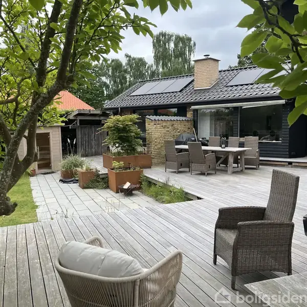 Terrasse med trædæk, hvor møbler er arrangeret til spisning. En have omkranser området med grønne planter og træer. Et moderne hus med solpaneler udgør baggrunden.