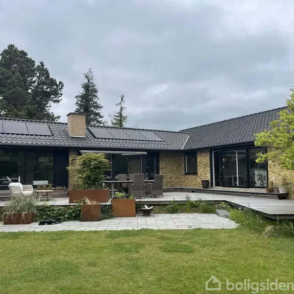 En moderne bungalow med solpaneler på taget, omgivet af en frodig have med græsplæne og træer. En terrasse med udendørs møbler strækker sig langs husets facade.