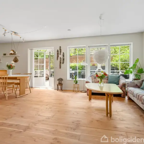 Stue med sofa og sofabord foran store vinduer, indeholder spisebord med stole. Udenfor ses grønne planter. Lyse vægge og gulve giver rummet et åbent og indbydende look.