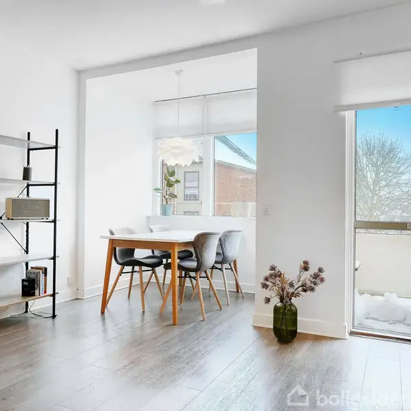 Et lyst stueområde med et sort reolsystem, et spisebord med fire stole, og en glasdør, der åbner ud til en altan med sne. Indretningen har moderne møbler og trægulv.