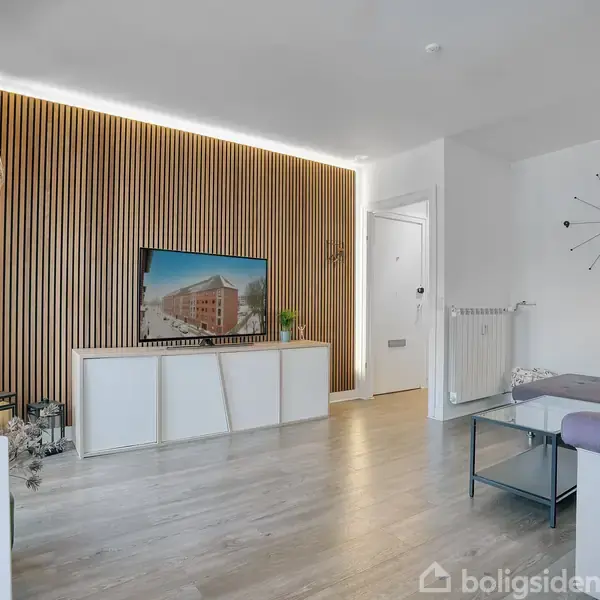 Stue med en L-formet sofa og glasbord; vægdekorationer og hængende lamper skaber hyggelig stemning. Hvidmalede vægge og lys trægulv supplerer moderne indretningen.