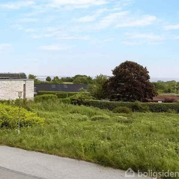 En græsbevokset tomt ligger stille ved en vej, omgivet af lavt buskads og en murstensbygning i et landligt område med spredte træer under en klar blå himmel.