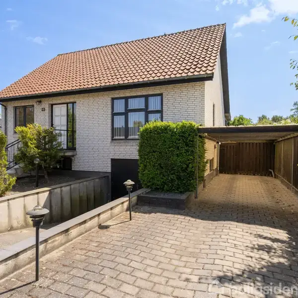 Huset står stille i en forstadshave med flisebelagt indkørsel og omkranset af velplejede buske og træer. Adressen 10 er synlig ved hoveddøren.