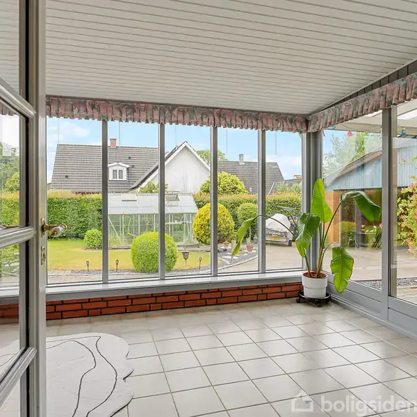 En udestue med flisegulv og store vinduespartier viser en velholdt have med buske og græsplæne udenfor. En grøn plante i en potte står ved siden af døren.