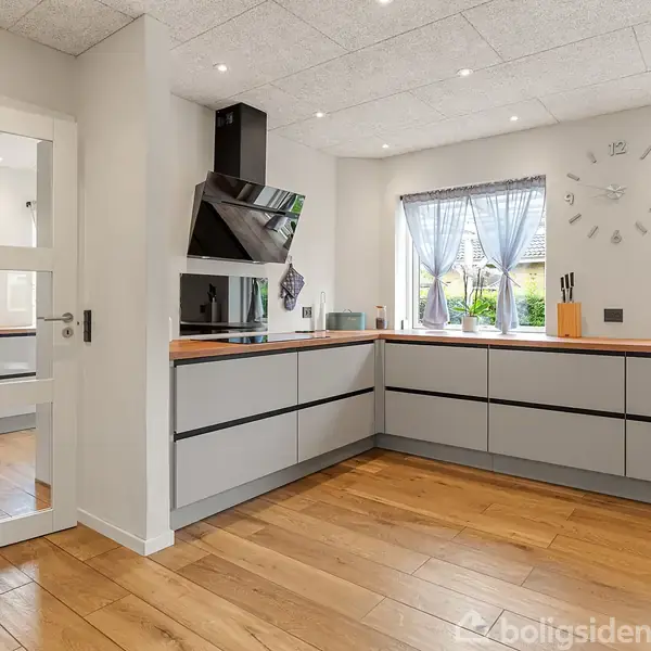 Køkkenet har lysegrå skabe og træbordplader, med en emhætte i hjørnet. To store vinduer med gardiner lader lys ind, og en vægur hænger på væggen. Gulvet er i lyst træ.