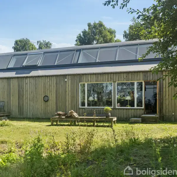 Et moderne træhus med store vinduer og solpaneler på taget ligger i en frodig have med græs og træer omkring.