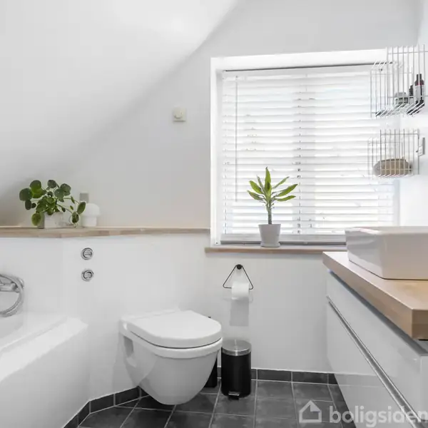Et lyst og moderne badeværelse med badekar, hængetoilet og vask forrest. En plante på vindueskarmen under persienner. Sorte fliser på gulvet. Vindueshylde har sammensatte genstande.