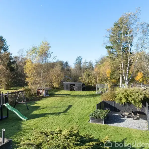 En grøn græsplæne med legepladsudstyr, inklusive en rutsjebane, omgivet af træer i en baghave. En terrasse med siddepladser findes til højre. Himlen er klar og blå.
