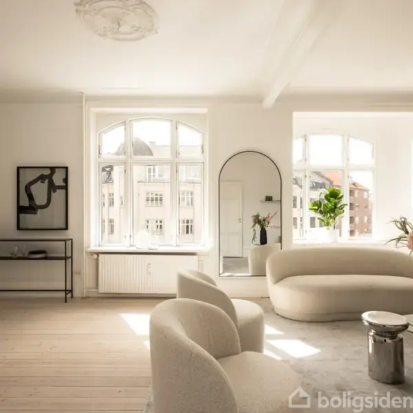 En lys stue med buede vinduer, en cremefarvet sofa, og stole. Et spejl står ved væggen. Der er planter og blomster i vaser. En minimalistisk indretning i moderne stil.