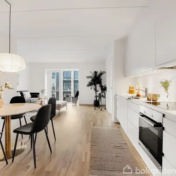 Et moderne køkken-alrum med hvide skabe og træbord, omgivet af spisestue og stue. Rummet er lyst med store vinduer og stilfuld indretning, herunder sorte stole og en lampe.