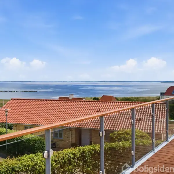 En terrasse med glasrækværk tilbyder udsigt over et roligt hav og rødt tag på en murstensbygning. Omkringliggende huse og grønne arealer ses i baggrunden under en blå himmel.