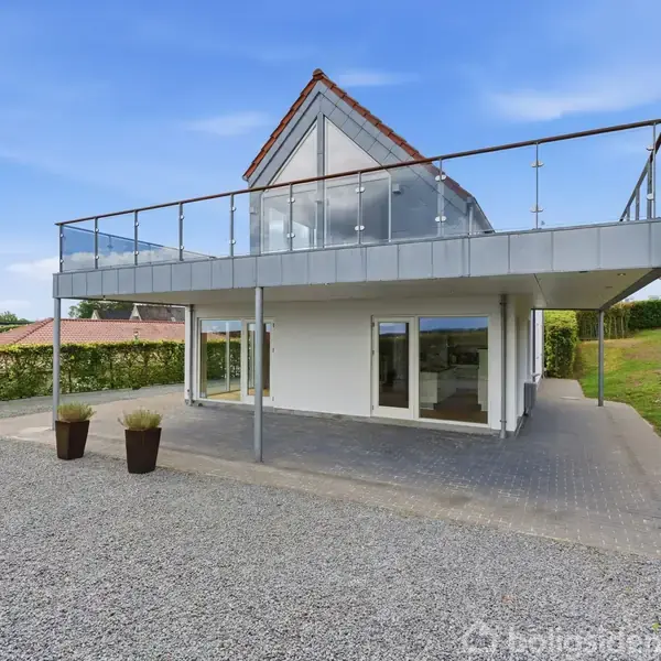 Et moderne hus med store vinduespartier og en øvre terrasse med glasrækværk står på en grusbelagt grund. Omkringliggende grønne områder skaber en fredelig atmosfære.