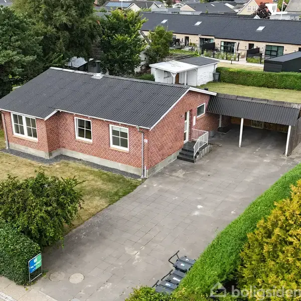 Et rødt murstenshus med sort tag står stille på en velplejet grund. En carport til højre og grønne hække omkranser haven. Tætliggende huse ses i baggrunden.