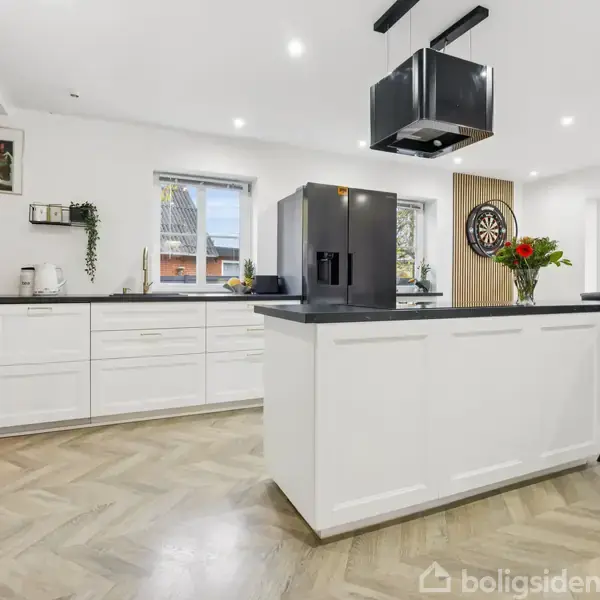 Moderne køkken med hvidt interiør og køkkenø, sort emhætte, og dobbeltdørs køleskab. To vinduer med udsigt til et tag. Blomstervase og dartskive pynter rummet. Ingen tekst synlig.