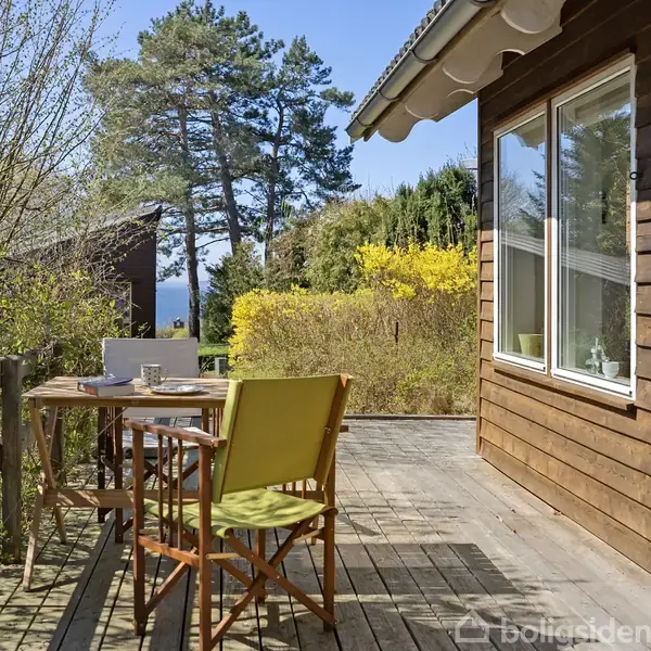 Træbord og -stole står stille på en træterrasse ved et brunt hus. Terrassen er omgivet af grønne buske og træer med blå himmel i baggrunden.