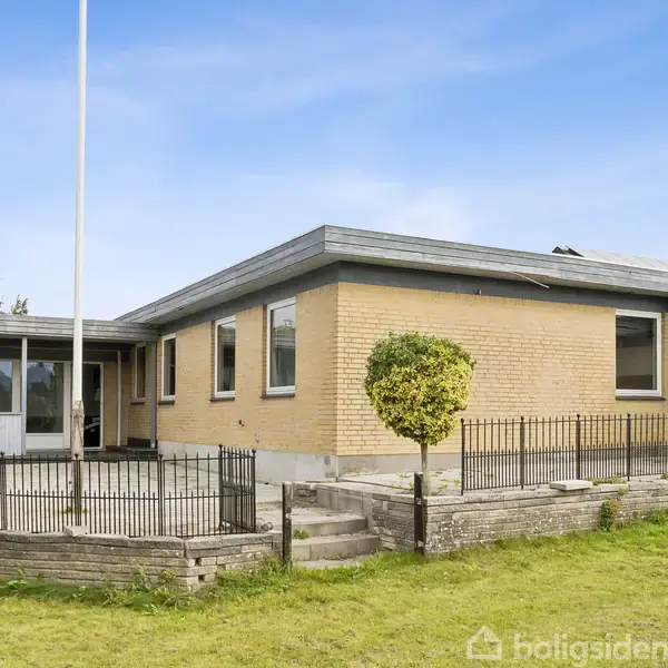Huset, en enkeltstående bygning i gule mursten, står stille i en velholdt have med grøn græsplæne og lavt hegn omkring terrassen. Himlen er klar og blå.