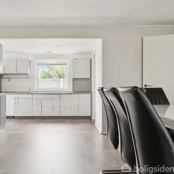 Et moderne køkken med hvide skabe; i forgrunden står sorte spisebordsstole ved et hvidt bord. Rummet er lyst, med et vindue, der viser grøn bevoksning udenfor.