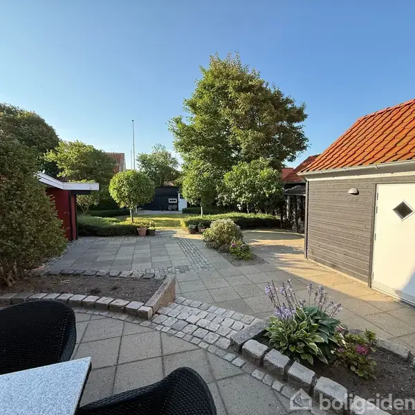 En brolagt gårdhave med en spiseplads har blomsterbede i forgrunden. Til venstre ses en rød murstensbygning, mens en garage med hvid dør er til højre. I baggrunden er grønne træer.