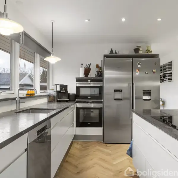 Et moderne køkken med grå bordplader og hvide skabe. Midtpunktet er et stort, rustfrit stål køleskab og indbyggede ovne. Rummet har trægulv, store vinduer og hængelamper.