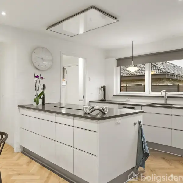 Køkken med hvidt skabsmiljø og stor køkkenø; en orkidé står på øen. Store vinduer udenfor viser et gråt tag. Væggen har et rundt ur. Ingen tekst.