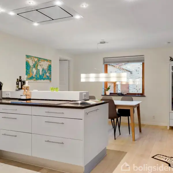 Køkken med hvide skabe og sort bordplade, forbundet til spiseområde med bord og stole. Væg med verdenskort-plakat. Glasskab til højre, vindue med udsigt til baghave.