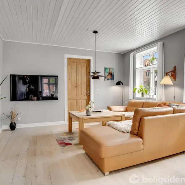 Brunt hjørnesofa, placeret i stue med lyse trævægge og loft. Tv hænger på væggen, planter og lamper dekorerer rummet. Store vinduer lader naturligt lys ind.