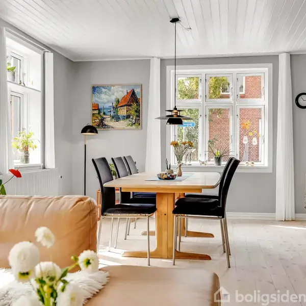 Stue med lædersofa står i forgrunden, mens et spisebord og stole er midt i rummet. Store vinduer lader lys strømme ind. En vægdekoration og planter skaber en varm atmosfære.