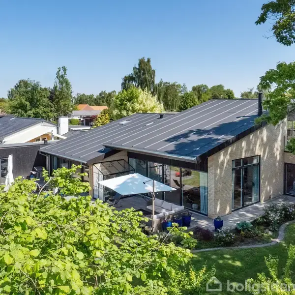 Stor moderne villa med skråt tag og store vinduer. Beliggende i frodig have med træer og terrasse. Omgivet af grønne områder og andre huse i et beboelsesområde.