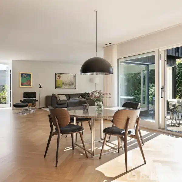Et moderne spisebord med fire stole står under en stor, sort lampe. Rummet er lyst og åbent med trægulv, sofa, lænestol og adgang til terrasse med udsigt til have.