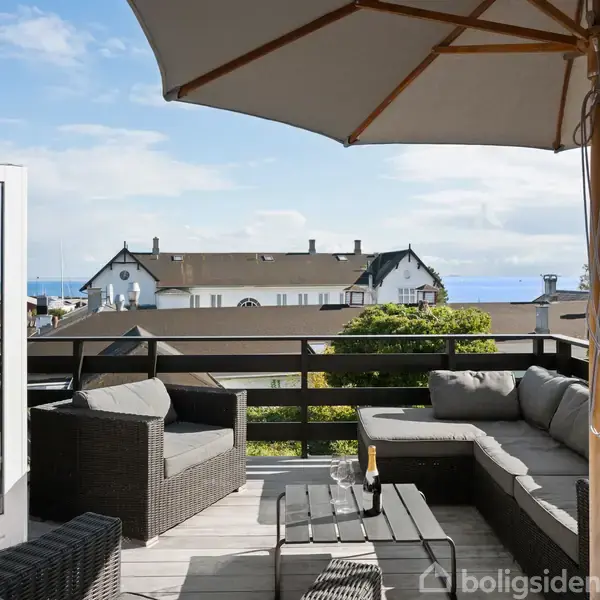 Terrassemøbler står under en parasol med udsigt over hustage og havet i baggrunden. Et glasbord og komfortable stole skaber en afslappet atmosfære i solskinsvejr.