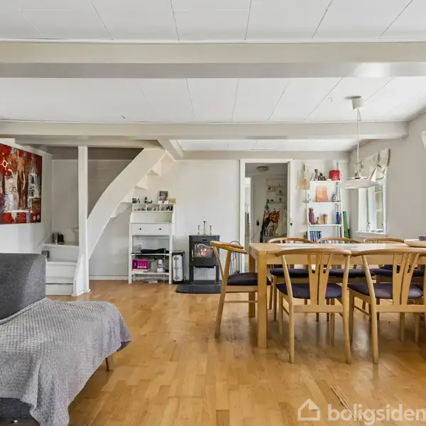 Stue med grå sofa og spisebord med seks stole, placeret på trægulv. Maleri og reol på væggen. Stort vindue giver udsigt til grøn natur udenfor.