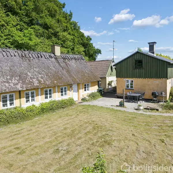 Et stråtækt hus står stille ved siden af en gul, to-etagers bygning med grøn gavl. Omgivet af klippet græsplæne og frodig skov under en blå himmel.