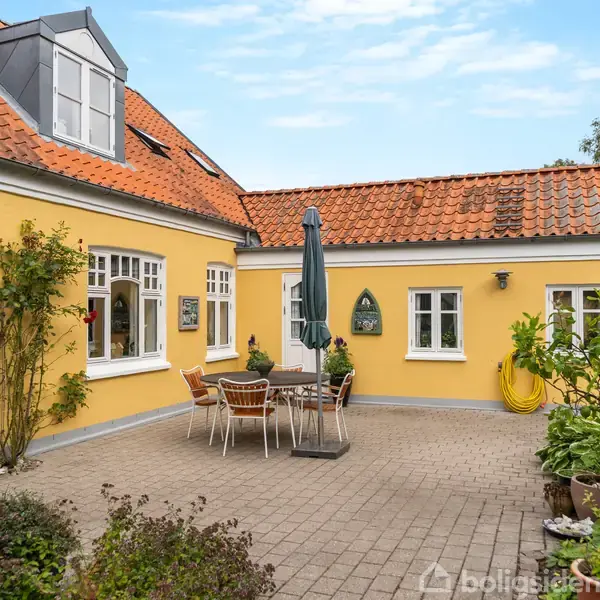 Gult hus omgivet af brostensbelagt gårdhave; bord og stole under en parasol i midten; planter og grønne områder omkring, med gule vægge og røde teglstenstage.