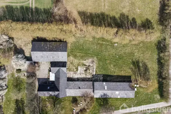 En landlig boligstruktur set oppefra står stille omgivet af frodige træer og grønne marker. Huset har flere tilsluttede bygninger og ligger ved en græsvej.