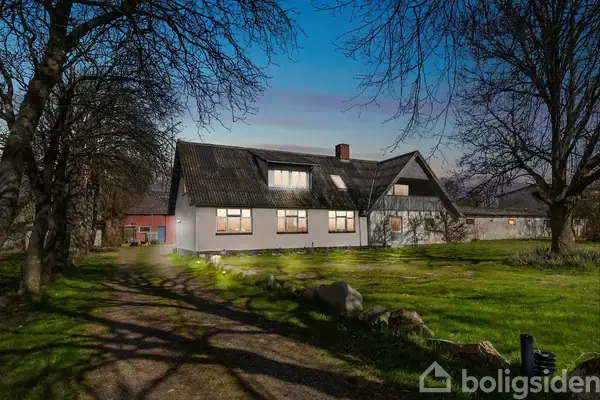 Huset står stille i skumringen med lys fra vinduerne, omgivet af en græsplæne med tætte træer og en grusvej, der fører op til indgangen.