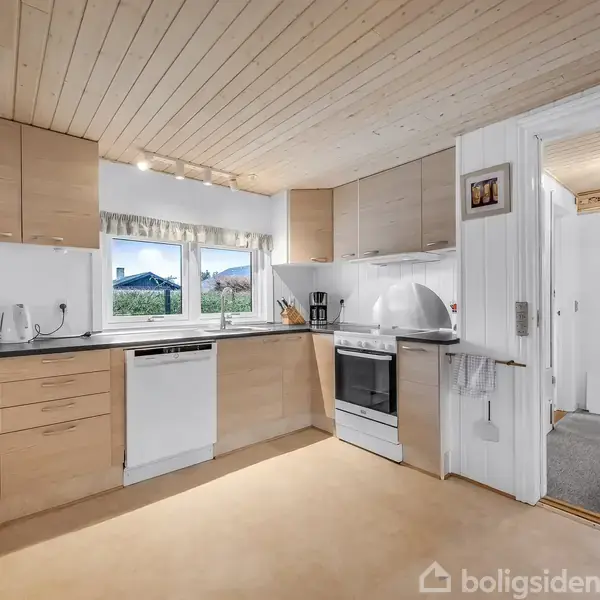 Køkken med lyse træskabe og sorte bordplader; en opvaskemaskine og komfur indbygget. Rummet er lyst med vinduer mod haven. Tilstødende gang leder til et andet rum.