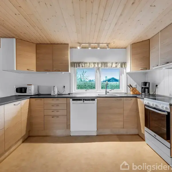 Et lyst køkken med træskabe og sort bordplade. Der står en elkedel, en kaffemaskine og en ovn. Udsigt til have gennem vinduet midt i rummet. Ingen tekst tilgængelig.