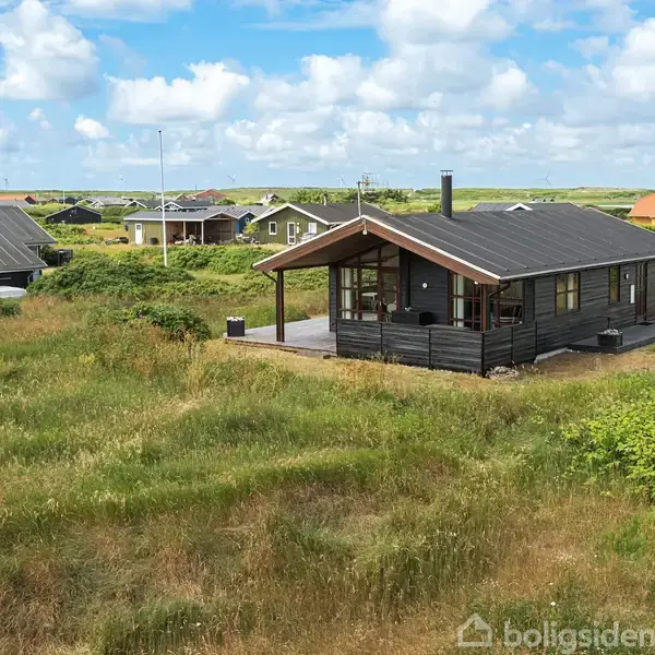 Et sort træhus står stille i en frodig, grøn mark med flere andre huse i baggrunden under en blå himmel med spredte skyer.
