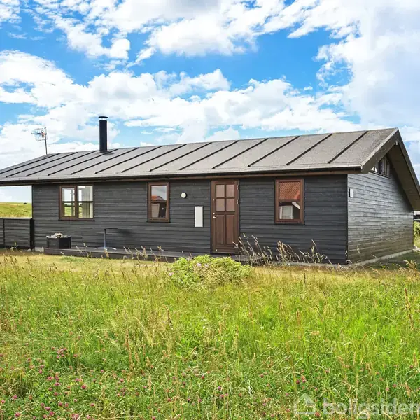 Mørkt træhus med skråt tag står i en åben græsmark. Huset har flere vinduer og en skorsten. Himlen er delvist skyet med blå pletter synlige.