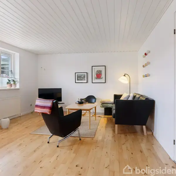 Stue med sort lænestol og sofa, placeret om et træbord. En lampe lyser op rummet med træloft og parketgulv. Store vinduer og planter skaber en lys og åben atmosfære.